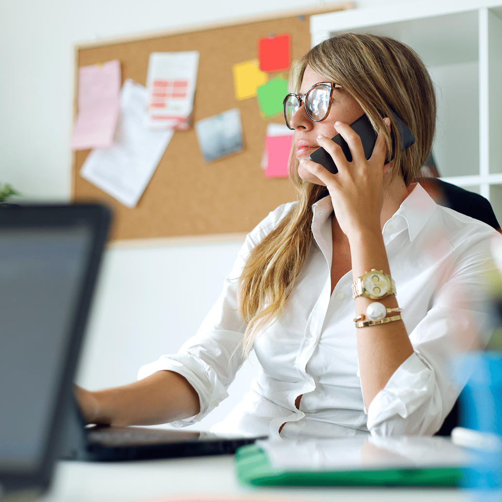 Woman wearing glasses sits at a desk in an office, talking on the phone while using a laptop, with a bulletin board and shelves in the background.